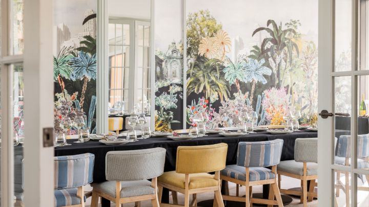 Pastel coloured chairs tucked into a long dining table in front of a tropical wall mural